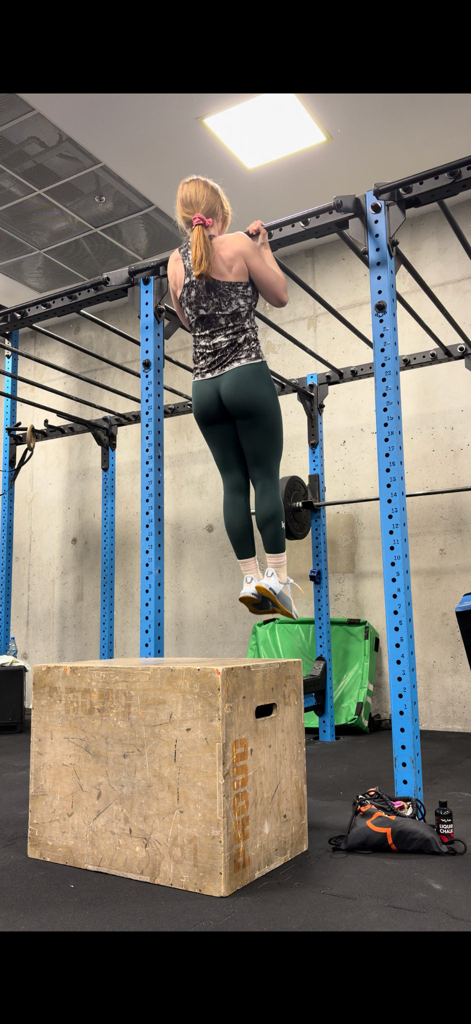 Kobieta w legginsach i topie wykonuje podciągnięcia na drążku w siłowni. Widoczny boks, sprzęt do ćwiczeń i betonowa ściana w tle. Oświetlenie sufitowe.