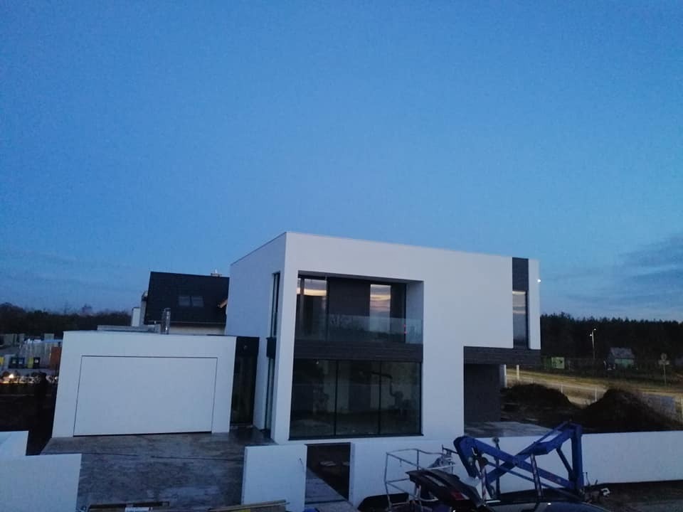 Nowoczesny dom z płaskim dachem i dużymi przeszkleniami, wykończony w bieli i szarości, sfotografowany o zmierzchu. Widoczne rusztowania i elementy budowlane.