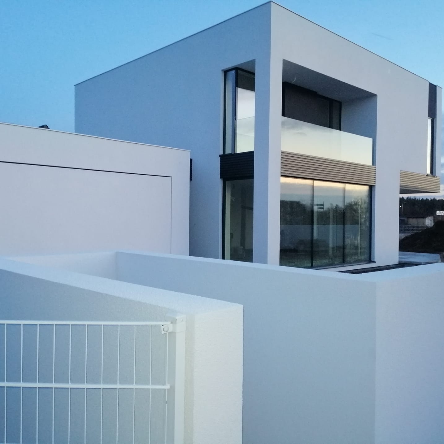 Nowoczesny, biały dom z płaskim dachem i dużymi oknami, widziany zza białego muru z metalowym ogrodzeniem. Minimalistyczna architektura w spokojnej, wieczornej scenerii.