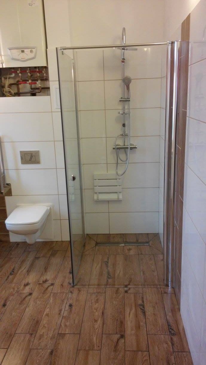 Nowoczesna kabina prysznicowa z siedziskiem i odpływem liniowym, wykończona płytkami imitującymi drewno. Na ścianie podwieszana toaleta i piec gazowy.