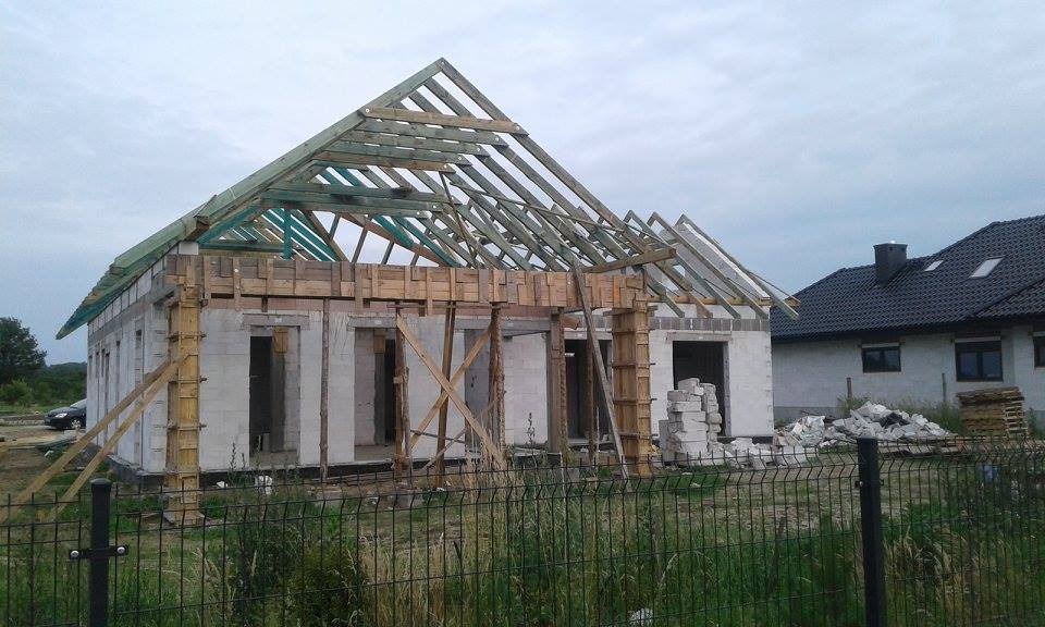 Budowa domu jednorodzinnego w stanie surowym otwartym: ściany z betonu komórkowego, drewniana konstrukcja dachu, szalunki. Widok zza ogrodzenia.