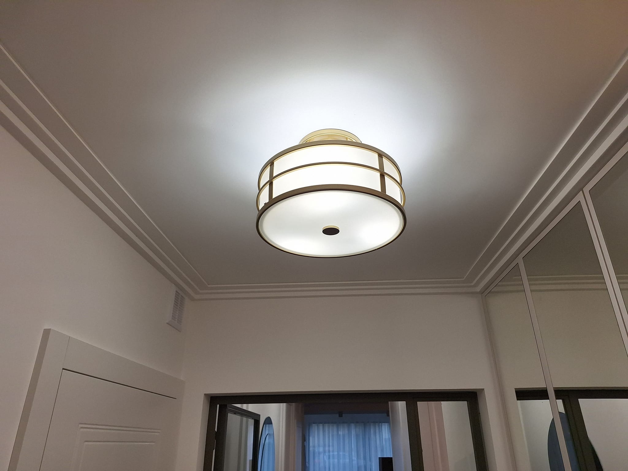 Nowoczesna lampa sufitowa z dekoracyjnymi listwami przysufitowymi w jasnym wnętrzu. Widoczne drzwi i fragment szafy z lustrami.