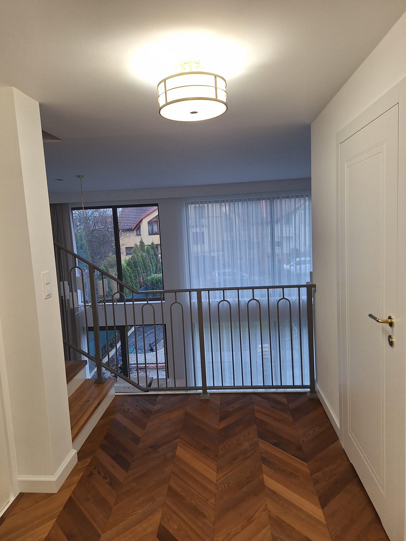 Parkiet w jodełkę, metalowa balustrada schodów i białe drzwi w jasnym korytarzu. Widok na zewnątrz przez okno z firankami. Nowoczesne oświetlenie sufitowe.