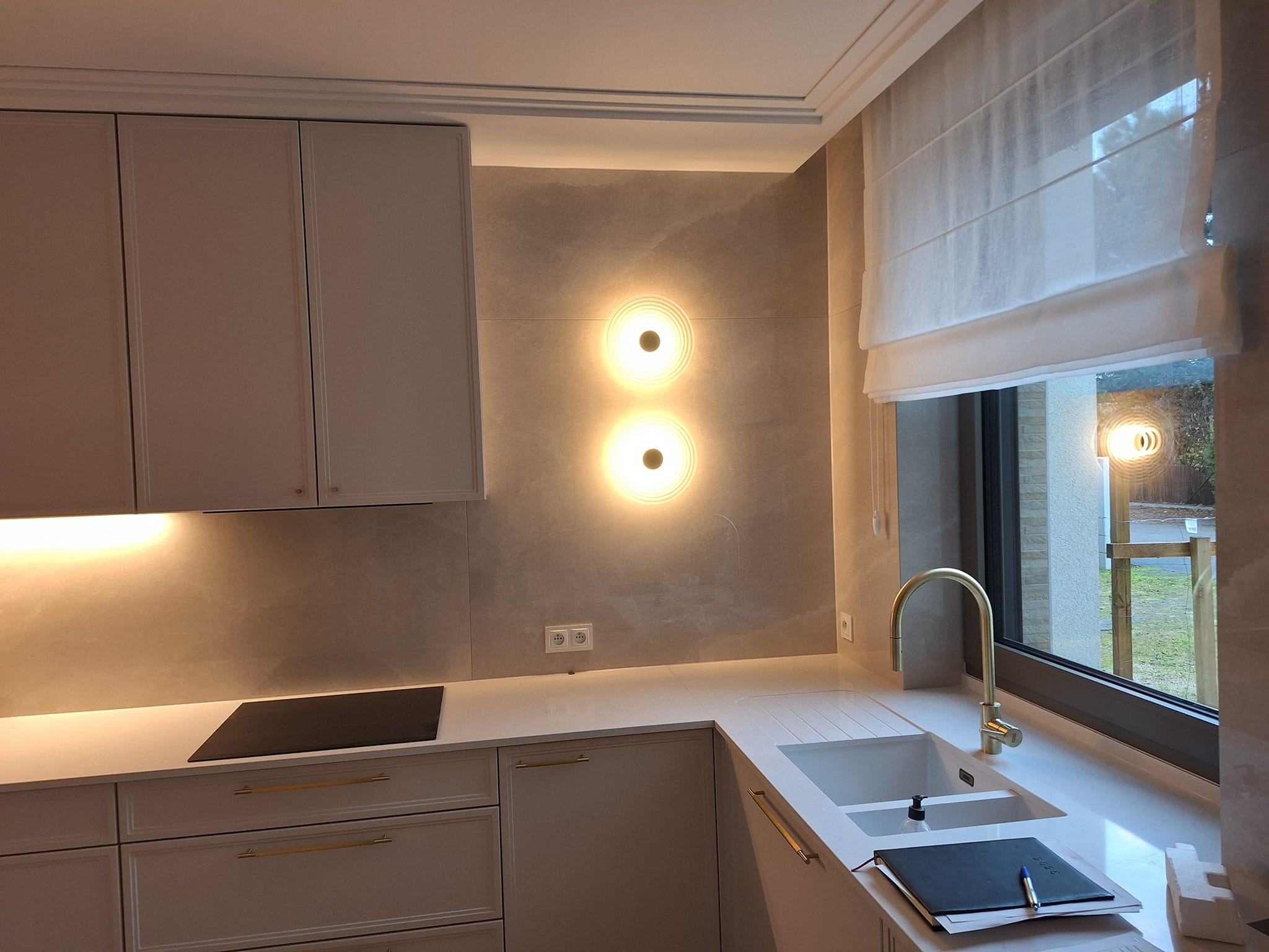 Elegancka kuchnia w odcieniach beżu z białymi szafkami, złotą armaturą i oświetleniem punktowym. Na blacie leży notatnik i długopis. Widok na zewnątrz przez okno.