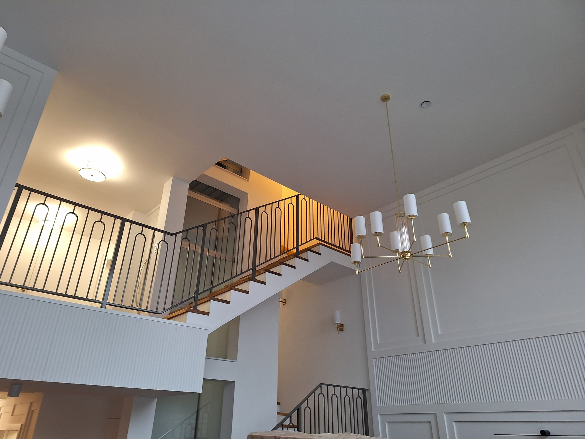 Nowoczesne schody z czarną balustradą i drewnianymi stopniami w jasnym wnętrzu. Na suficie designerski, złoty żyrandol. Ściany zdobione sztukaterią.