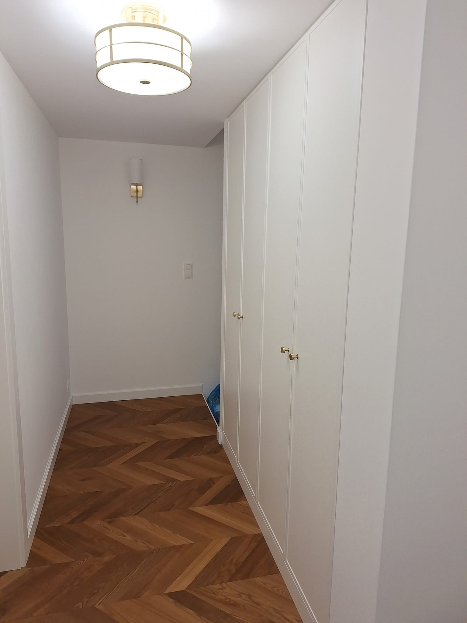 Korytarz z parkietem w jodełkę i białą zabudową szafy z złotymi uchwytami. Na ścianie kinkiet i włącznik. Oświetlenie sufitowe w stylu retro.