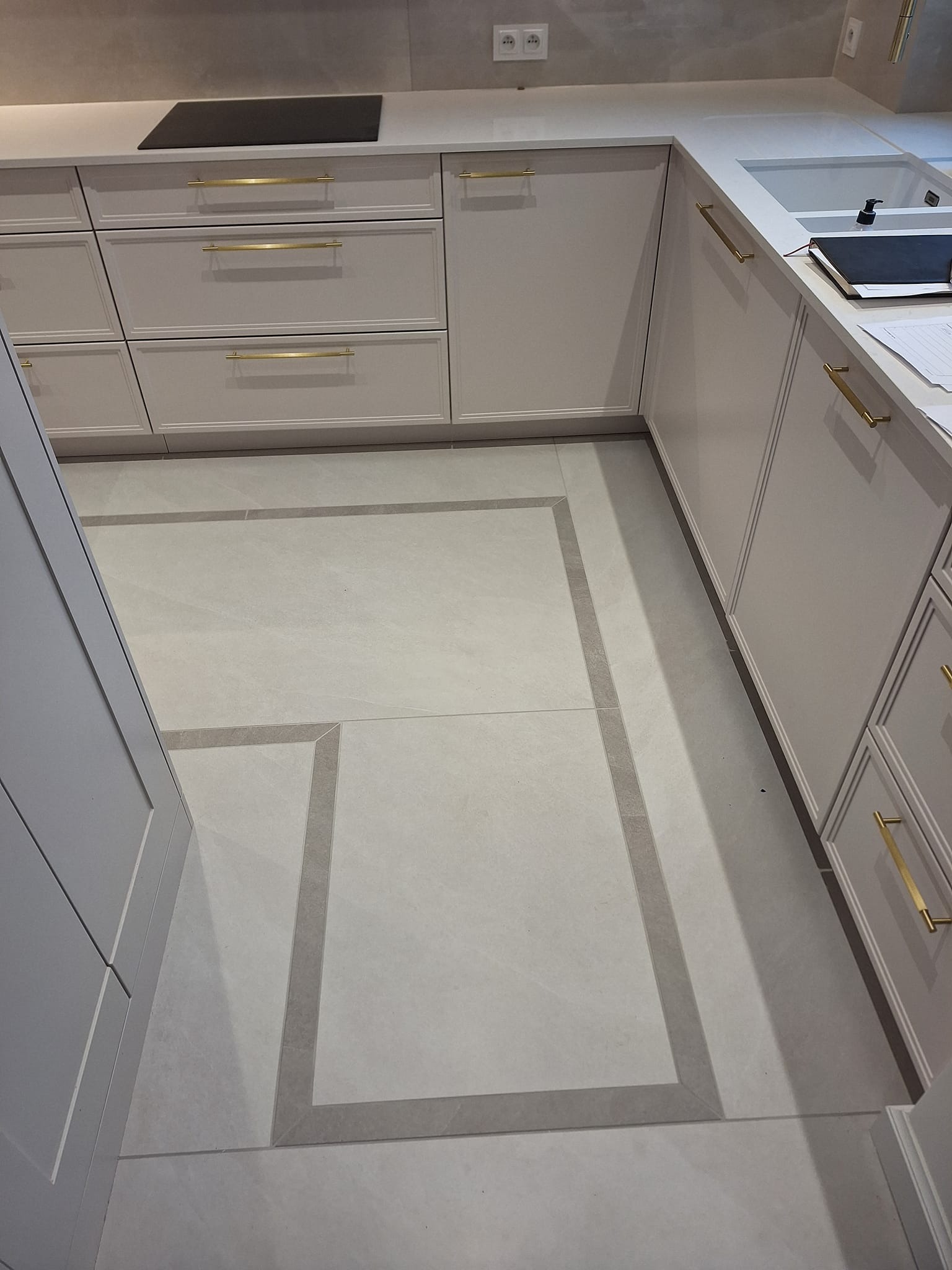 Jasna kuchnia z białymi szafkami i złotymi uchwytami. Na podłodze jasne płytki z szarym obramowaniem. Widoczne gniazdka elektryczne i fragment blatu z akcesoriami.