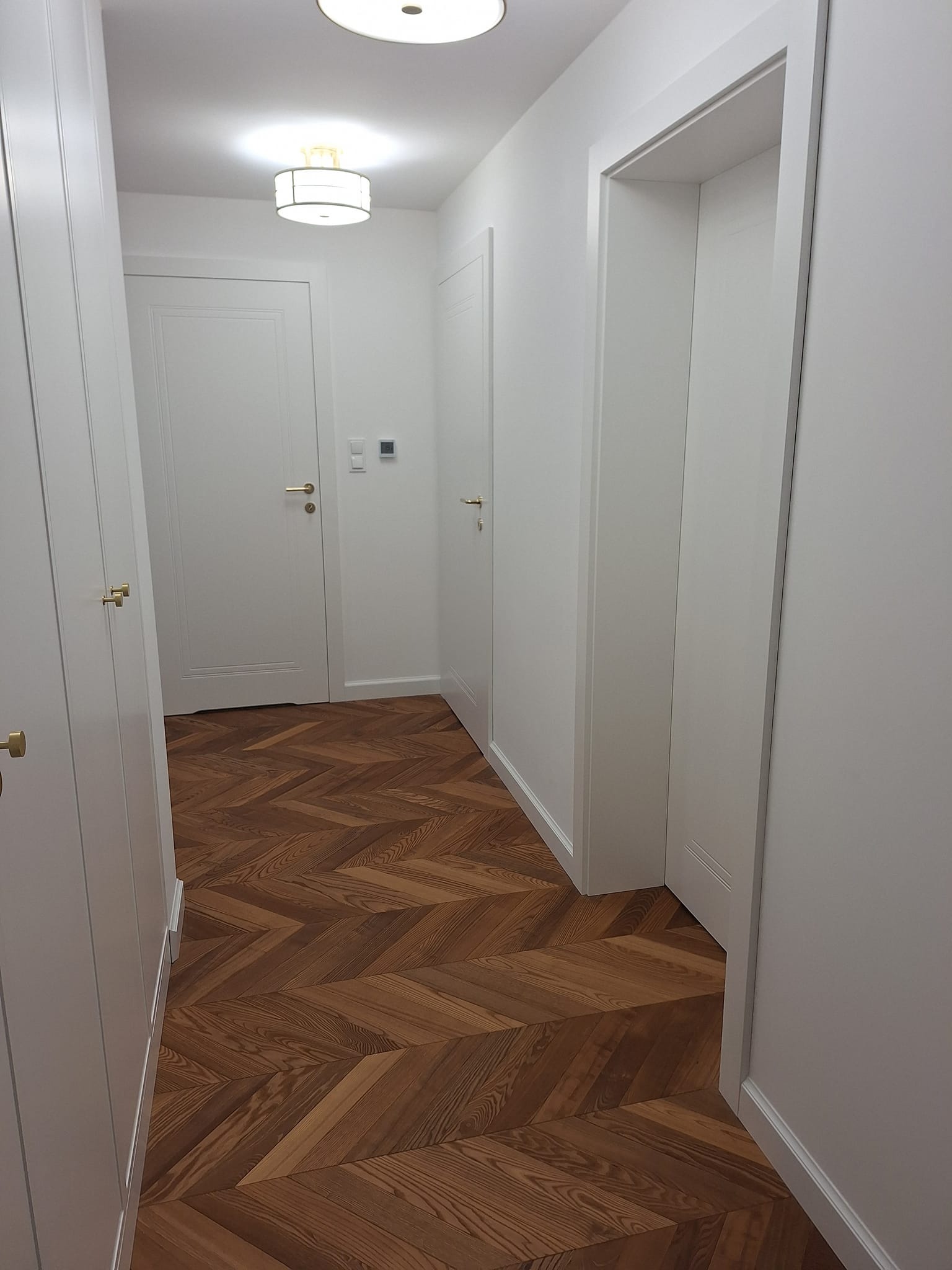 Białe drzwi i szafy w korytarzu z parkietem ułożonym we wzór jodełki. Złote okucia dodają elegancji. Oświetlenie sufitowe.