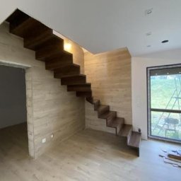 Projekt-Schody - Nowoczesne, drewniane schody wiszące w jasnym wnętrzu domu w Łomiankach. Minimalistyczny design, drewno w ciepłym odcieniu, widok z dołu na konstrukcję.