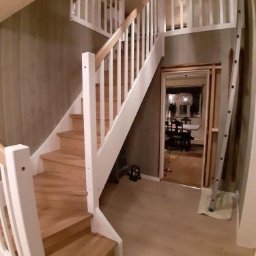 Projekt-Schody - Jasne drewniane schody z białymi balustradami, widok z dołu. W tle widoczne wnętrze domu i drabina oparta o ścianę. Oświetlenie sufitowe w stylu nowoczesnym.