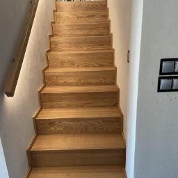 Projekt-Schody - Jasne, dębowe schody bez balustrad, oświetlone listwą LED, prowadzące w górę. Na podeście widoczne dekoracyjne lampiony. Minimalistyczny design, naturalne wykończenie.