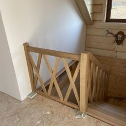 Projekt-Schody - Jasna, drewniana balustrada schodowa z krzyżowym wzorem, w surowym wnętrzu z boazerią i oknem. Widoczne schody i fragment ściany.