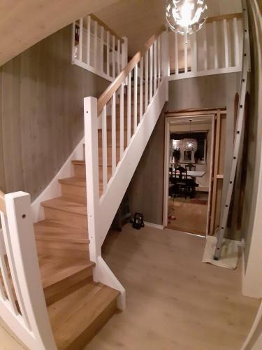 Jasne drewniane schody z białymi balustradami, widok z dołu. W tle widoczne wnętrze domu i drabina oparta o ścianę. Oświetlenie sufitowe w stylu nowoczesnym.