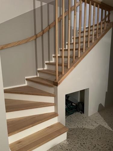 Jasne schody drewniane z prostą balustradą z okrągłych belek, zintegrowane ze ścianą, z widocznym schowkiem pod spodem i posadzką lastryko.