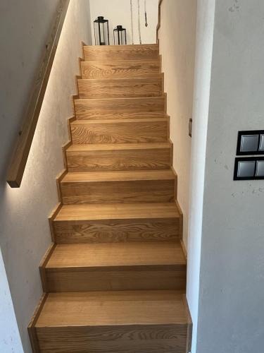 Jasne, dębowe schody bez balustrad, oświetlone listwą LED, prowadzące w górę. Na podeście widoczne dekoracyjne lampiony. Minimalistyczny design, naturalne wykończenie.