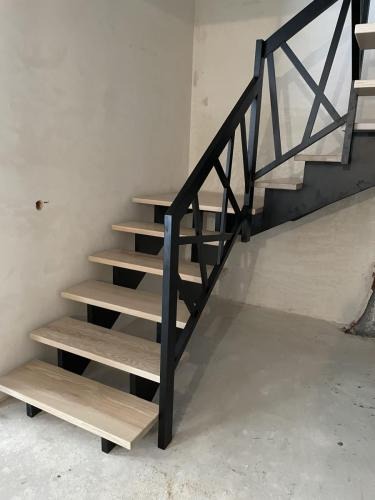 Schody z jasnego drewna z czarną, metalową balustradą o geometrycznym wzorze krzyży. Widok z dołu, surowy beton i ściany w tle.