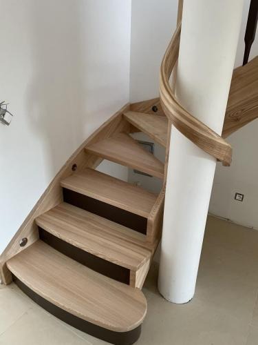 Spiralne schody drewniane z jasnego drewna, z ciemniejszymi podstopnicami, wsparte na białej kolumnie. Naturalne oświetlenie podkreśla detale.