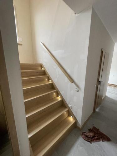 Jasne schody drewniane z podświetleniem LED na stopniach, z drewnianą poręczą przy ścianie. Widok z dołu, w tle fragment drzwi i podłogi.