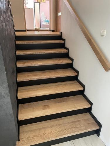 Nowoczesne schody z drewnianymi stopniami i czarnymi podstopnicami, oświetlone naturalnym światłem, z drewnianą poręczą przy białej ścianie.