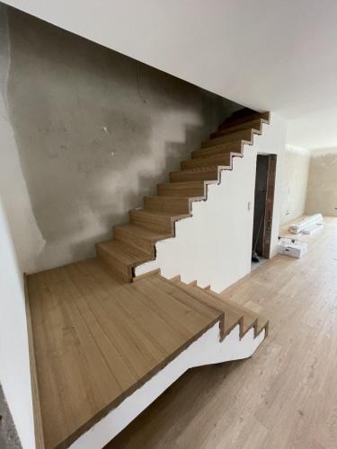 Montaż drewnianych schodów w surowym wnętrzu. Widoczne niedokończone ściany i podłoga. Jasne drewno kontrastuje z szarością betonu.