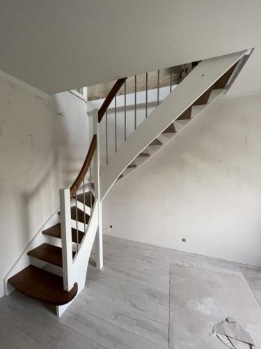 Klatka schodowa w trakcie montażu: drewniane stopnie, białe boki i metalowe pręty balustrady. Widoczne niedokończone ściany i podłoga.