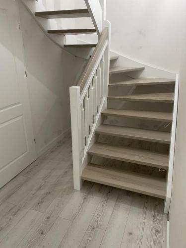 Drewniane schody z białą balustradą w jasnym wnętrzu. Widoczne drewniane stopnie i białe elementy konstrukcyjne. Podłoga wyłożona panelami imitującymi drewno.