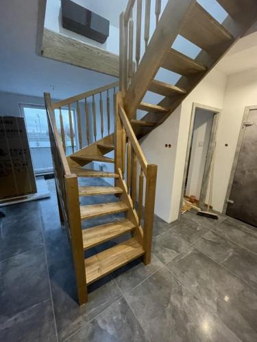 Drewniane schody spiralne z jasnego drewna, z balustradą z pionowych szczebli, na tle szarej podłogi z płytek. Widok z dołu, częściowo widoczne okno i drzwi.