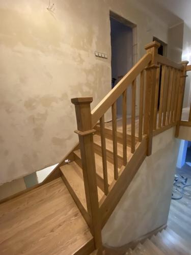 Jasne, drewniane schody z balustradą w surowym wnętrzu. Widoczne ślady po pracach wykończeniowych na ścianach. Naturalne oświetlenie podkreśla strukturę drewna.