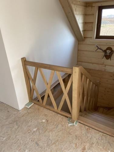 Jasna, drewniana balustrada schodowa z krzyżowym wzorem, w surowym wnętrzu z boazerią i oknem. Widoczne schody i fragment ściany.