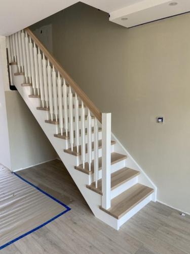 Jasne schody z drewnianymi stopniami i białą balustradą w nowoczesnym wnętrzu. Widoczne zabezpieczenia podłogi folią malarską.