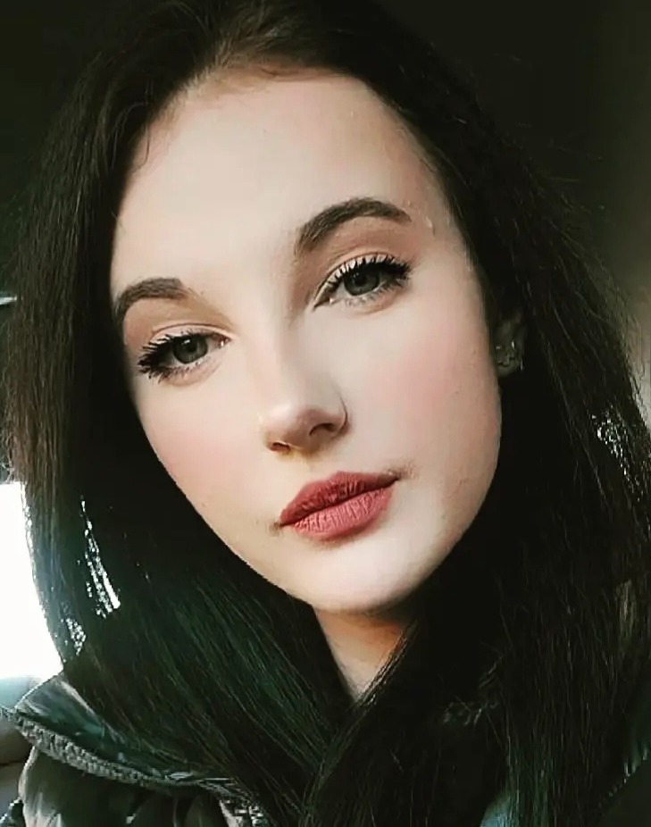 Portret młodej kobiety z ciemnymi włosami i delikatnym makijażem, skierowanej w stronę obiektywu, z subtelnym uśmiechem i naturalnym oświetleniem.