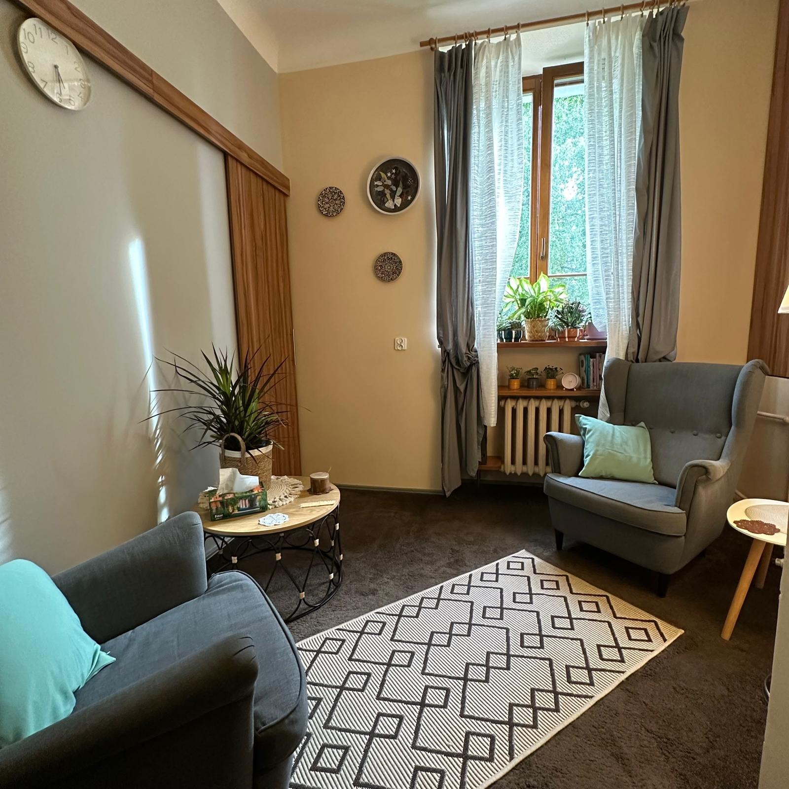 Przytulny gabinet z dwoma fotelami, dywanem w geometryczne wzory i roślinami w oknie. Na ścianie zegar i dekoracyjne talerze. Stonowana kolorystyka.