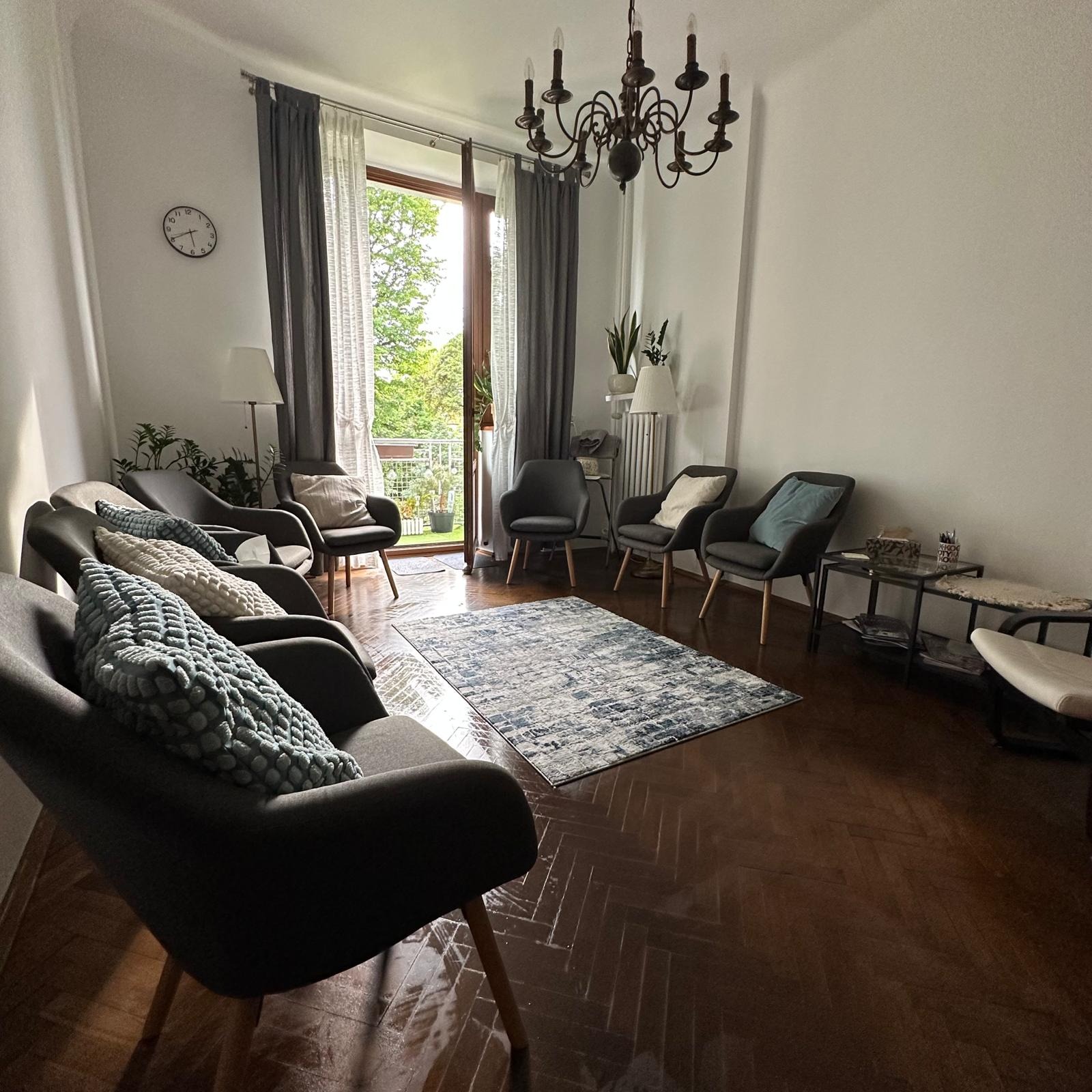 Jasne wnętrze gabinetu z krzesłami dla pacjentów, dywanem i balkonem z widokiem na zieleń. Na ścianie zegar, a nad krzesłami wisi ozdobny żyrandol.
