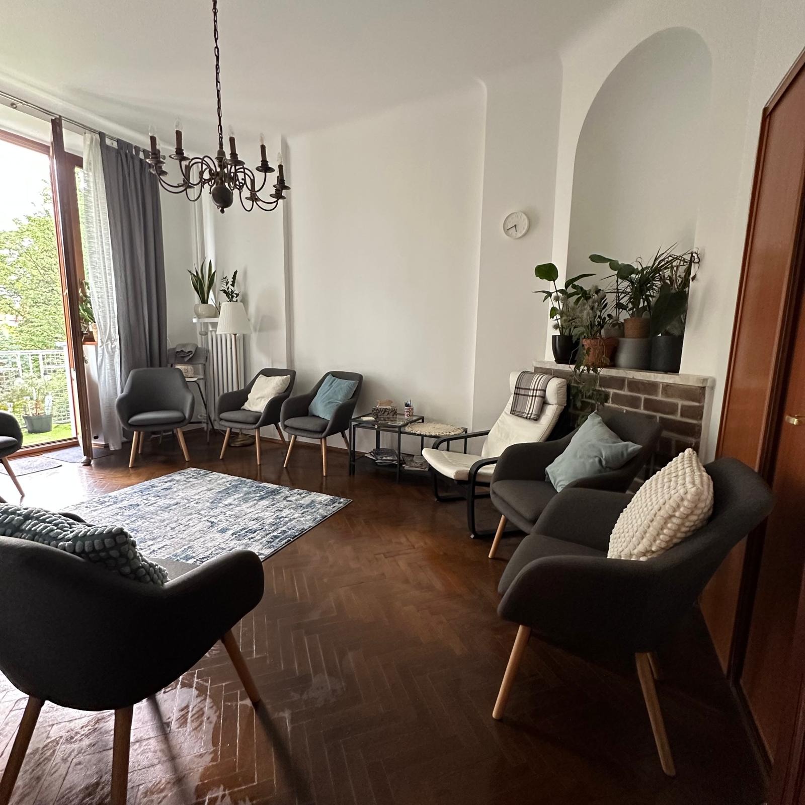 Jasne wnętrze gabinetu z krzesłami ustawionymi w kręgu, dywanem w geometryczne wzory i roślinami doniczkowymi. Widoczny parkiet i żyrandol.