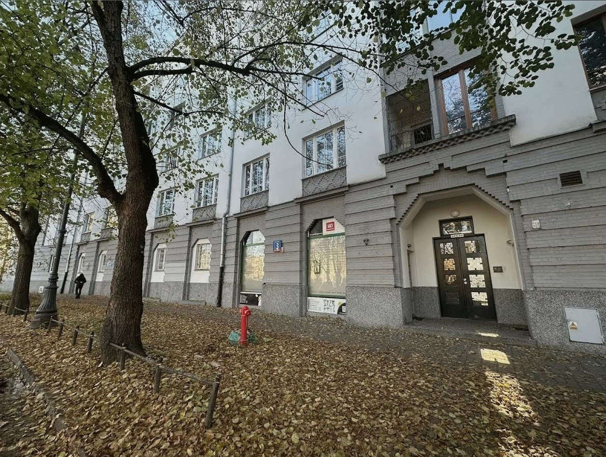 Wejście do budynku z ciemnymi drzwiami, widoczne z zewnątrz, z opadłymi liśćmi na chodniku i drzewami po lewej stronie. Tabliczka z numerem 3 na ścianie.