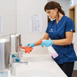 Fresh Office - Kobieta w niebieskim uniformie, w błękitnych rękawiczkach, czyści umywalkę pomarańczową ściereczką i spryskiwaczem w łazience z białymi płytkami i instrukcją mycia rąk.