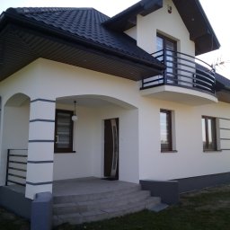 DAN-BUD Daniel Jenoch - Nowoczesny dom jednorodzinny z białymi ścianami elewacyjnymi, ciemną dachówką i elementami dekoracyjnymi w postaci poziomych pasów na kolumnach werandy, balkon z czarną balustradą, widoczny...