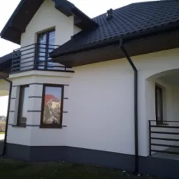 Nowoczesny dom jednorodzinny z białymi ścianami i ciemną dachówką, widoczny balkon z metalową balustradą, okna z brązowymi ramami, szara podmurówka i fragment trawnika.