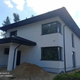 Nowoczesny dom z jasną elewacją i ciemną stolarką okienną, widok z niskiej perspektywy, z widoczną stertą ziemi przed budynkiem.