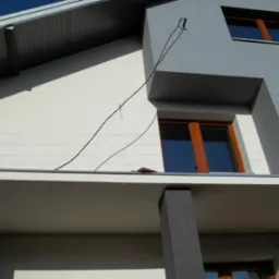 Dwukolorowa elewacja domu z drewnianą stolarką okienną i widocznym dachem. Szara kolumna podtrzymuje balkon. Widok z dołu, błękitne niebo w tle.