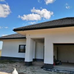 DAN-BUD Daniel Jenoch - Nowoczesny dom jednorodzinny z białymi ścianami i ciemną dachówką, widok z zewnątrz, w tle błękitne niebo z chmurami, teren wokół domu częściowo zagospodarowany, widoczne ślady prac budowlanych.