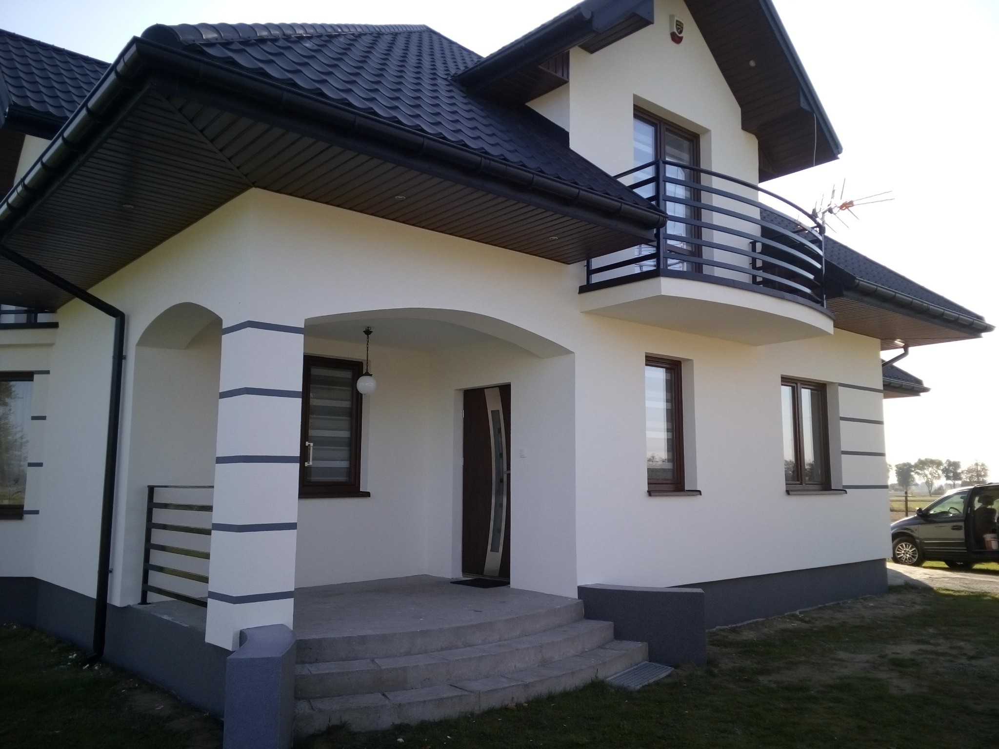 Nowoczesny dom jednorodzinny z białymi ścianami elewacyjnymi, ciemną dachówką i elementami dekoracyjnymi w postaci poziomych pasów na kolumnach werandy, balkon z czarną balustradą, widoczny...