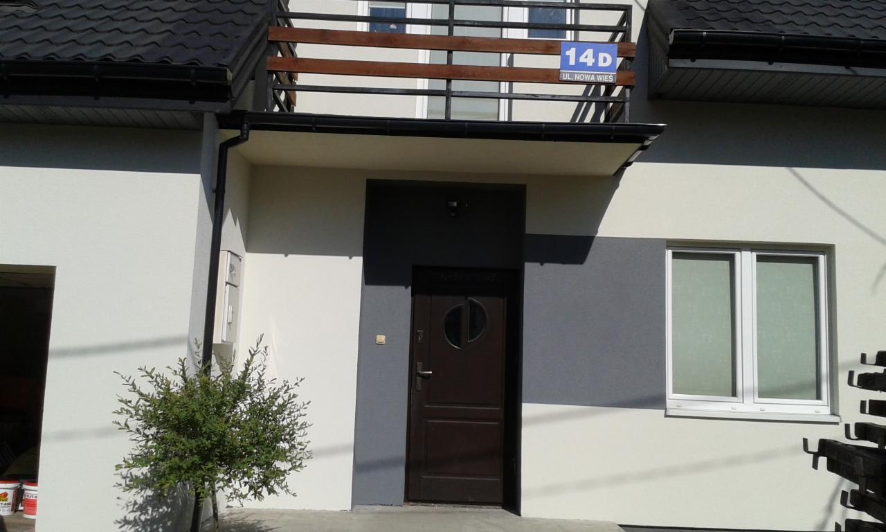 Elewacja domu jednorodzinnego z brązowymi drzwiami wejściowymi i oknem, fragment balkonu z drewnianą balustradą, adres 14D na tabliczce.