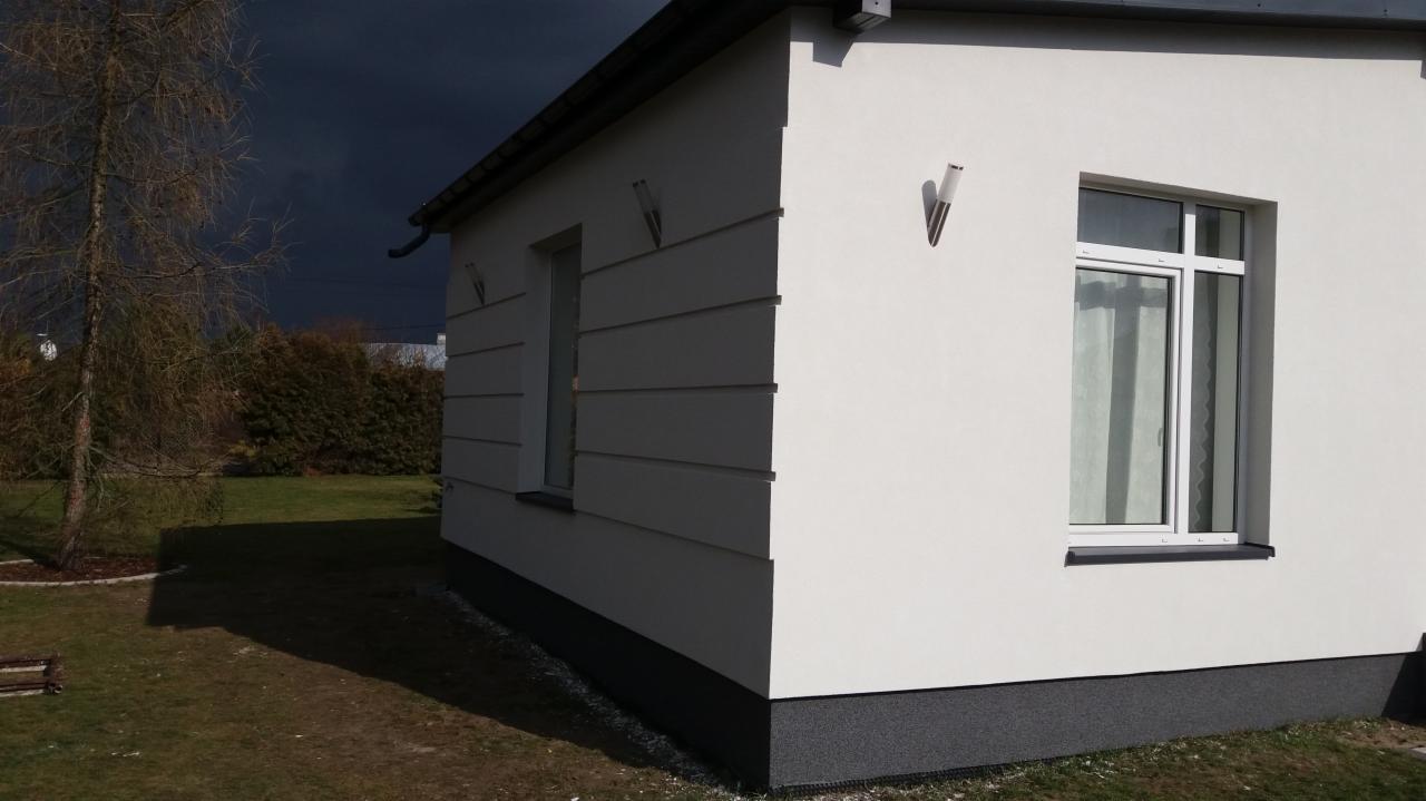 Nowoczesny dom jednorodzinny z białą elewacją i szarym cokołem, widoczne lampy elewacyjne w kształcie stożków, na tle ciemnego, burzowego nieba.