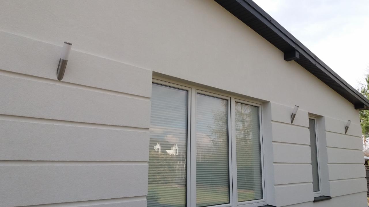 Fragment elewacji domu jednorodzinnego z białym tynkiem strukturalnym i ozdobnymi listwami, oświetlony dwoma kinkietami.