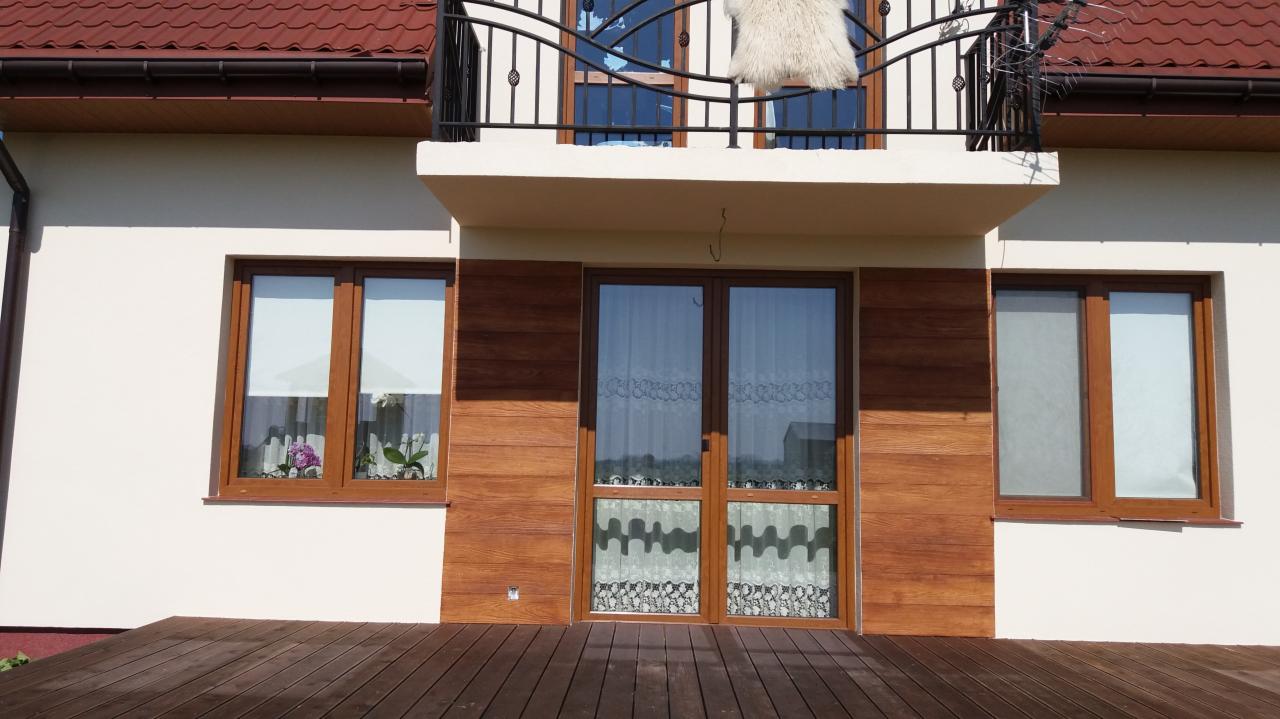 Elewacja domu jednorodzinnego z drewnianą okładziną wokół okien i drzwi balkonowych, balkon z czarną balustradą, brązowy dach, widoczny fragment drewnianego tarasu.