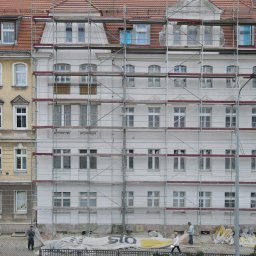 PARKER Budownictwo - Budynek w rusztowaniach, część fasady pomalowana na biało, druga w oryginalnym kolorze. Widoczni przechodnie i materiały budowlane.