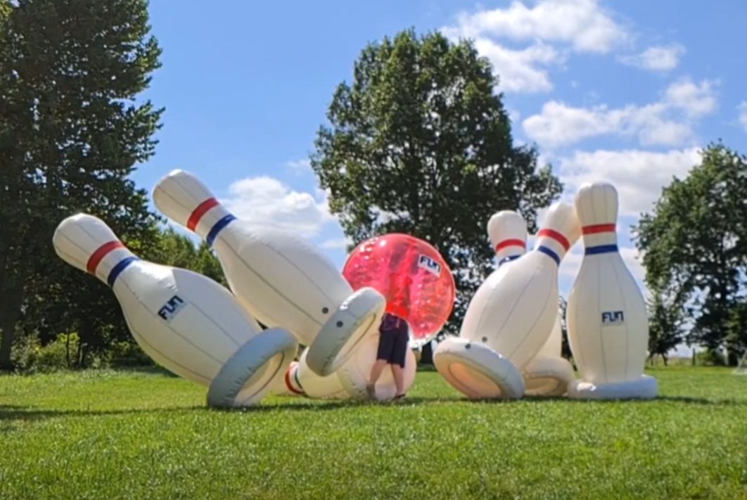 Dmuchane kręgle z osobą w kuli na trawie, w tle drzewa i błękitne niebo. Atrakcja eventowa z logiem 'FUN' na kręglach.