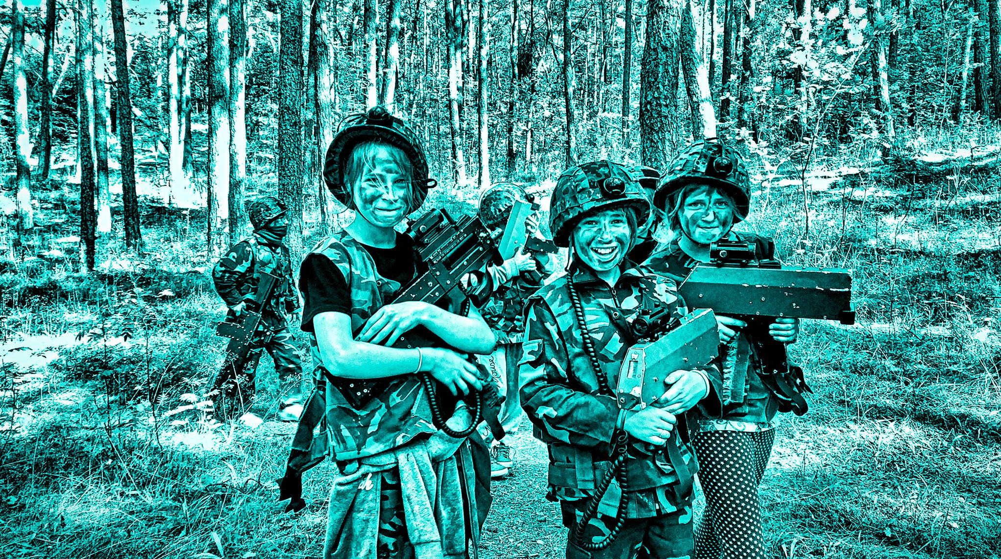 Grupa dzieci w lesie, w strojach militarnych i z laserowymi pistoletami, uśmiechnięte, z pomalowanymi twarzami, w trakcie zabawy w paintball laserowy.