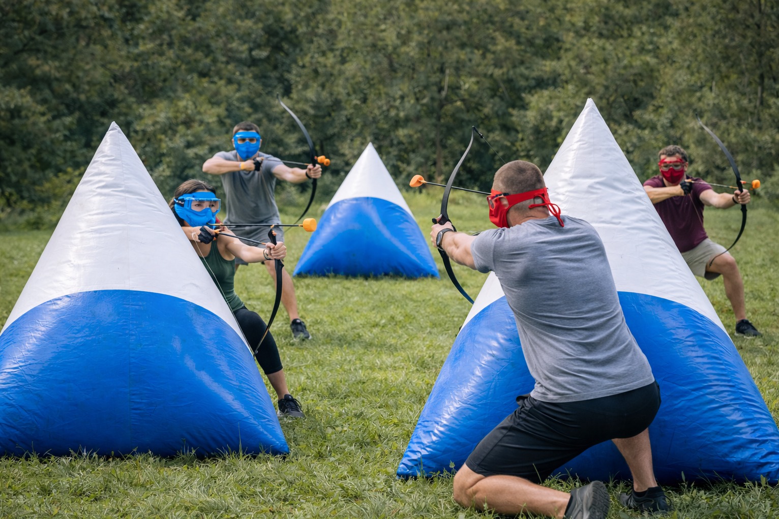 Gra w Archery Tag: grupa osób w maskach celuje z łuków za dmuchanymi przeszkodami na trawiastym terenie. Dynamiczne ujęcie akcji, widoczne napięcie łuków i skupienie graczy.